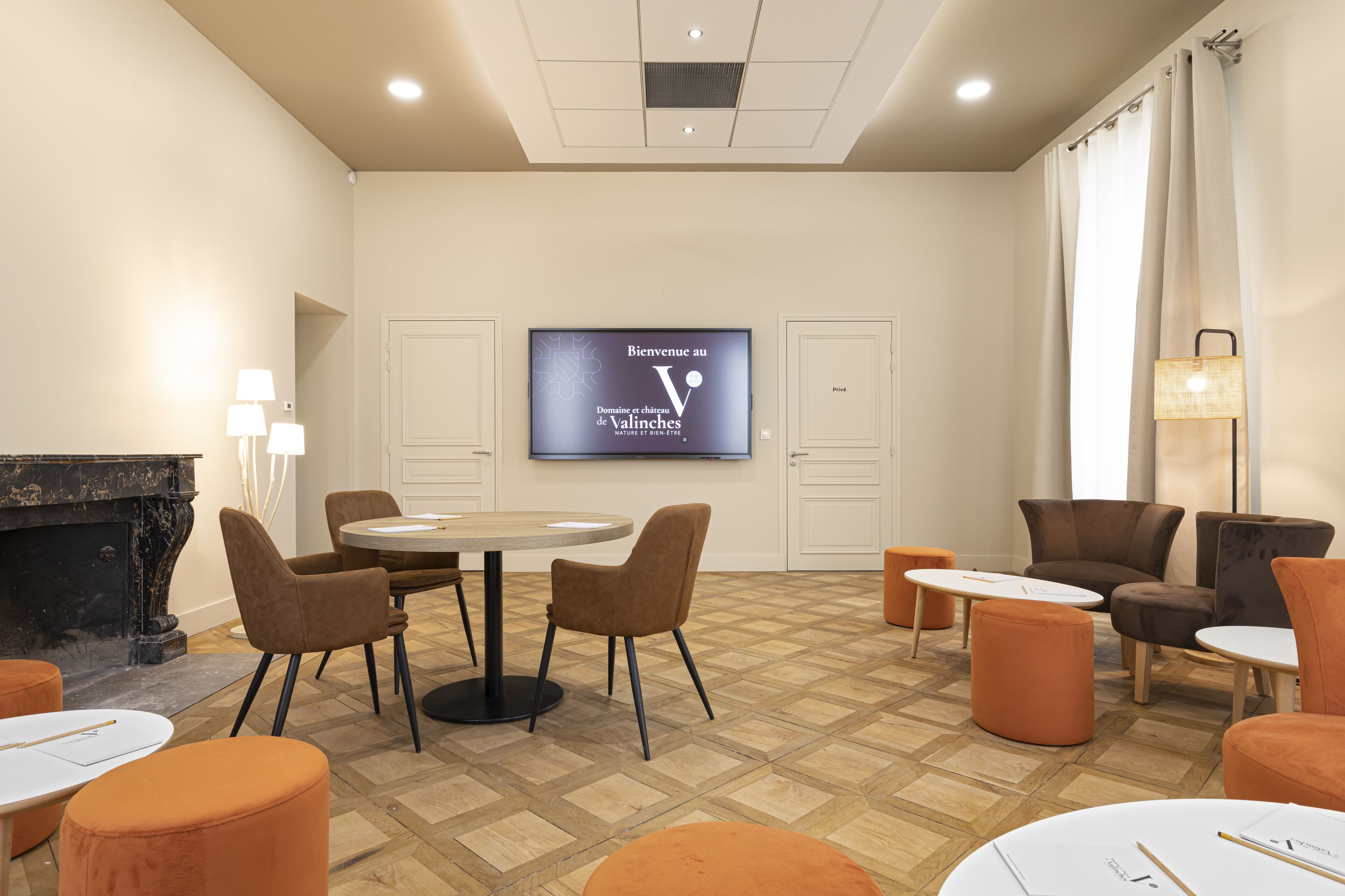 A modern hotel room with a fireplace, a TV screen displaying a welcome message, and a seating area with ottomans and chairs.
