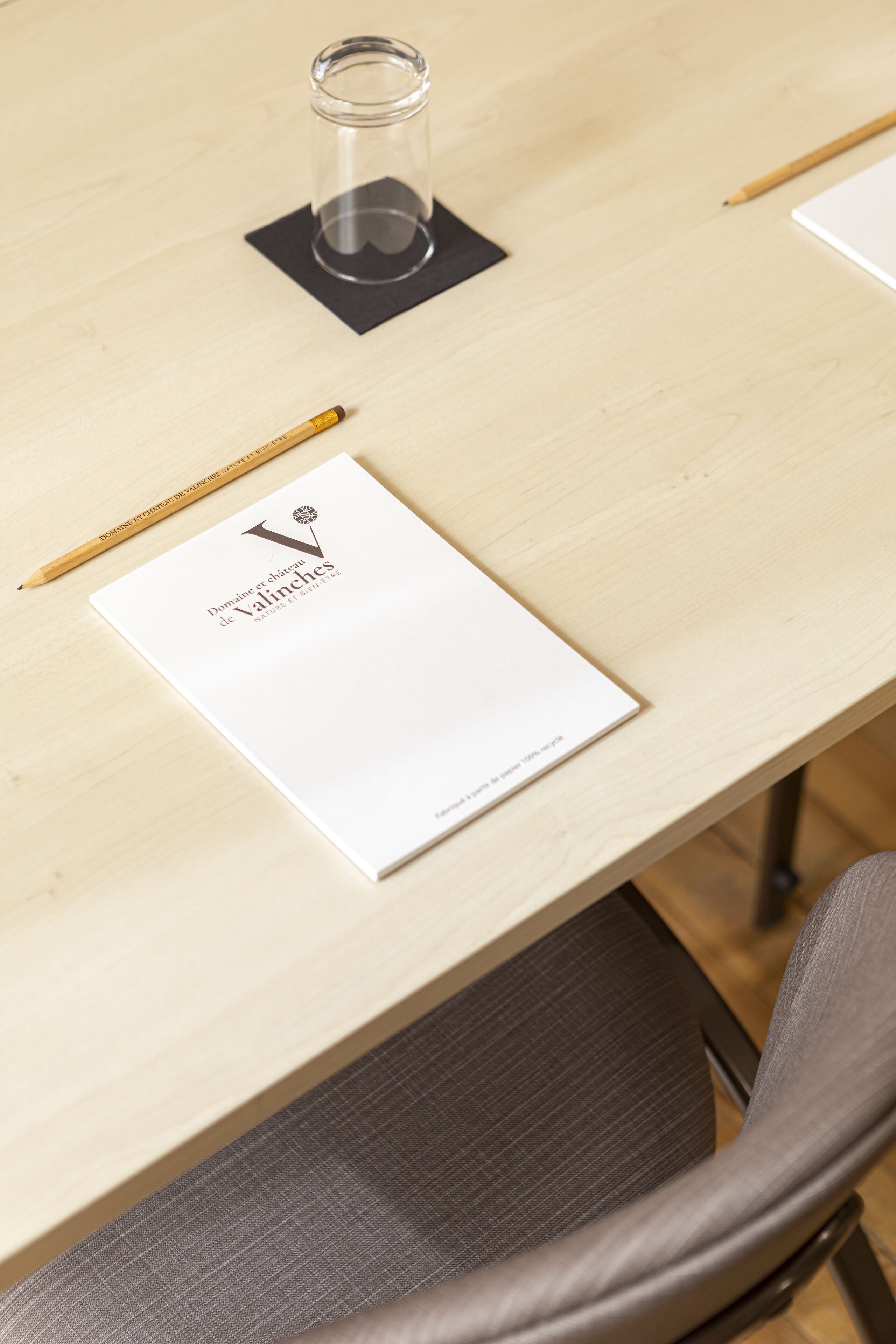 Una mesa de madera con un folleto blanco titulado 'Domaine Château de Valinches', un lápiz y un vaso sobre un posavasos negro.