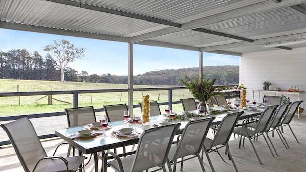 An outdoor dining area with tables set for a meal, chairs, and a view of a grassy field.