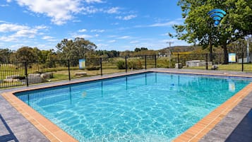 A rectangular swimming pool with a tiled edge, surrounded by a black metal fence, and a grassy area with trees in the background.