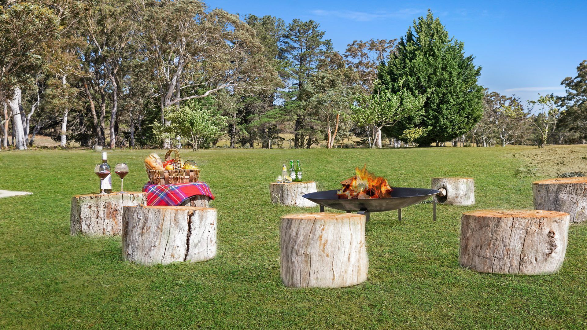 An outdoor setting with a picnic setup on a grassy field, featuring a fire pit, wooden stools, and a picnic basket.