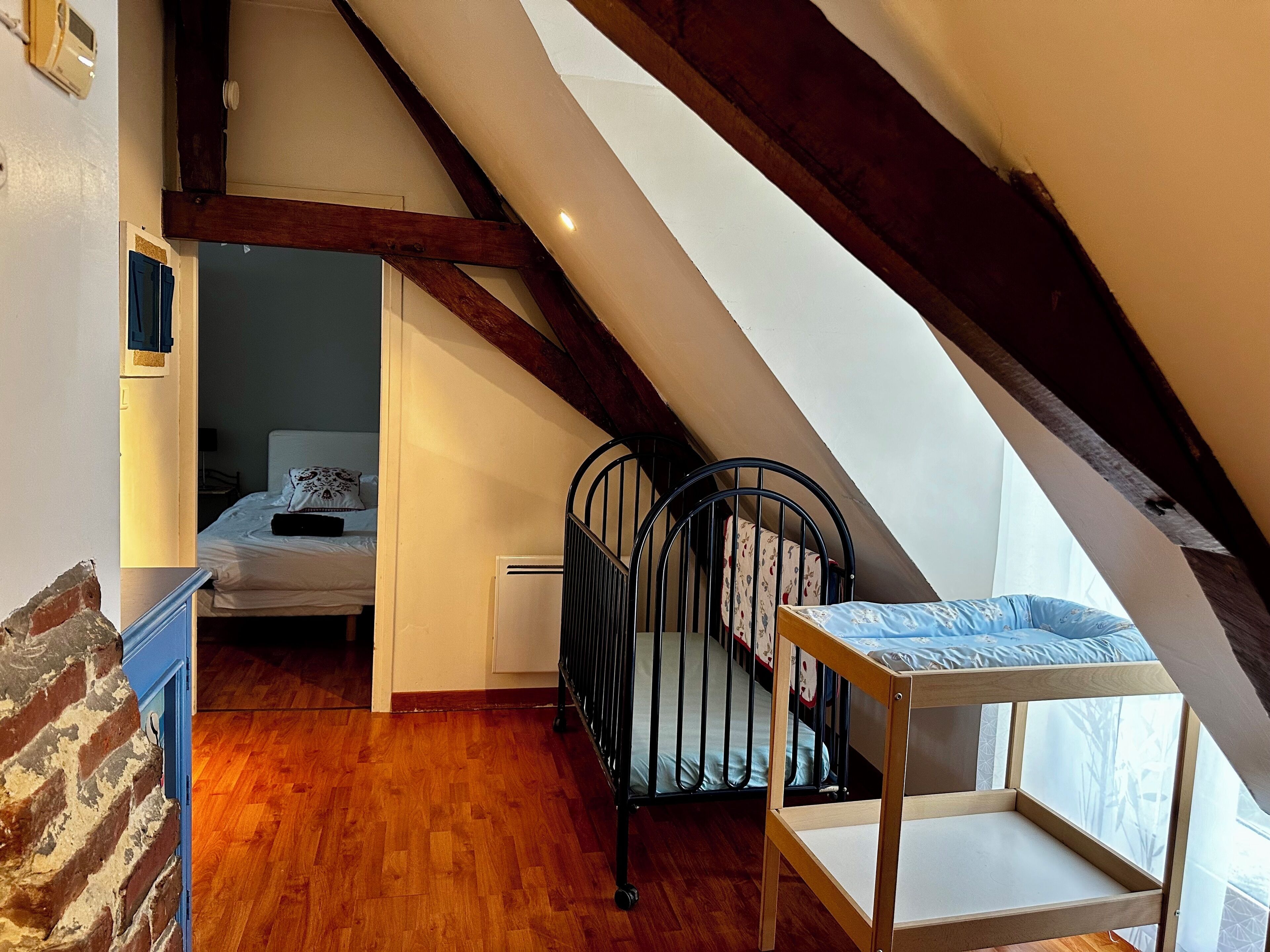 A room with a sloped ceiling, wooden beams, a bed, a crib, and a small table.