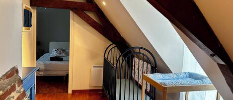 A room with a sloped ceiling, wooden beams, a bed, a crib, and a small table.