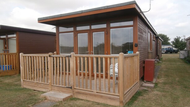 Une cabane en bois dotée d’une terrasse entourée d’une clôture, de grandes fenêtres et d’un petit porche.