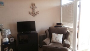 Un salon avec un téléviseur posé sur un meuble, une ancre décorative accrochée au mur, une fenêtre avec des stores et un fauteuil marron agrémenté d’un coussin.