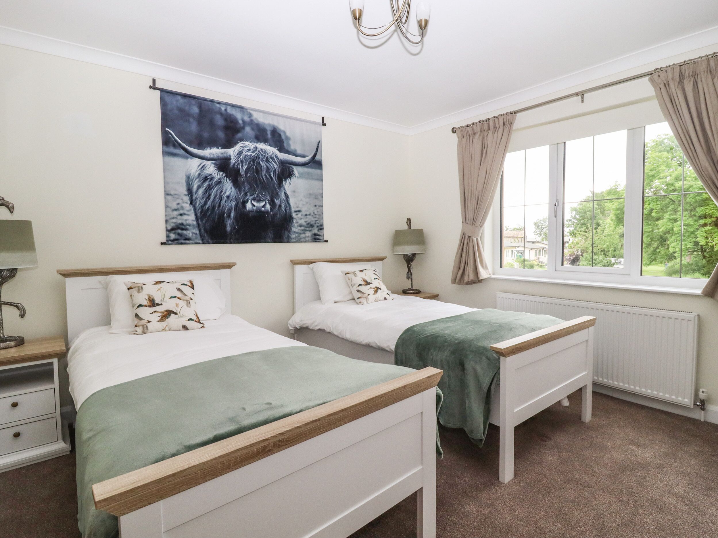 A hotel room with two single beds, a large window with curtains, a chandelier, and a wall-mounted artwork of a bull.