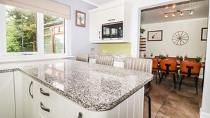 A modern kitchen with granite countertops, white cabinetry, and a built-in oven. There is a dining area with a wooden table and chairs, and a large window providing a view of the outdoors. - Bowland Fell Cottage (Skipton)