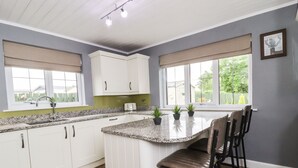 A kitchen with white cabinets, a granite countertop, a built-in island with bar stools, and a window with a view of greenery. - Bowland Fell Cottage (Skipton)