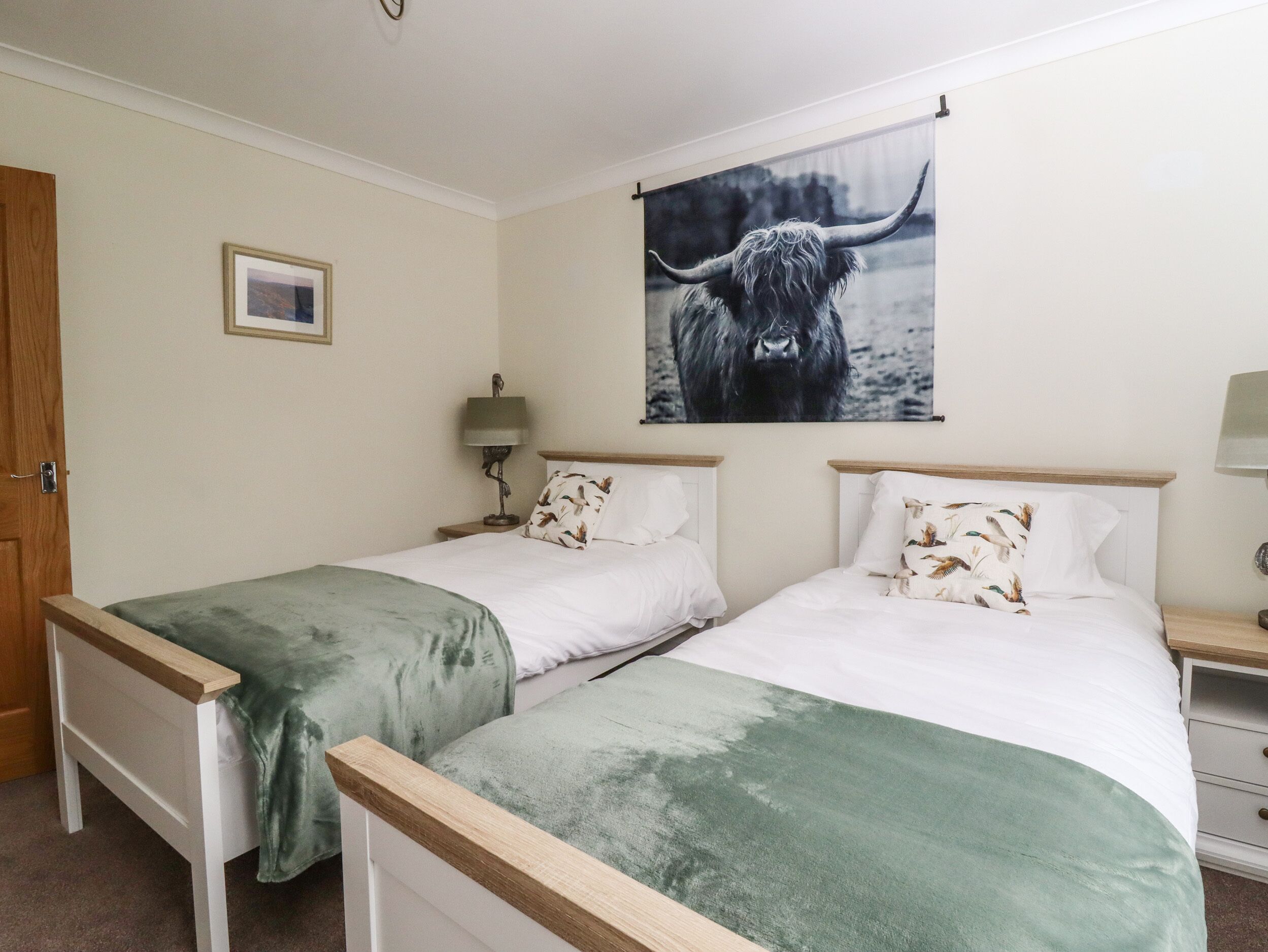 A hotel room with two single beds, a wooden headboard, a green blanket, a framed picture on the wall, and a lamp on the nightstand.