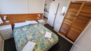 A bedroom with a wooden headboard, a bed with a colorful patterned bedspread, a nightstand, a wardrobe, and a small kitchenette in the background. - Close to Beach, Harbour Marina, Country Club, Golf Course, Restaurants, Cafes (Bermagui)
