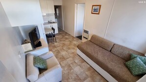 A living room with a sofa, a television, and a small table. There is a kitchen area with cabinets and a sink in the background. - Close to Beach, Harbour Marina, Country Club, Golf Course, Restaurants, Cafes (Bermagui)