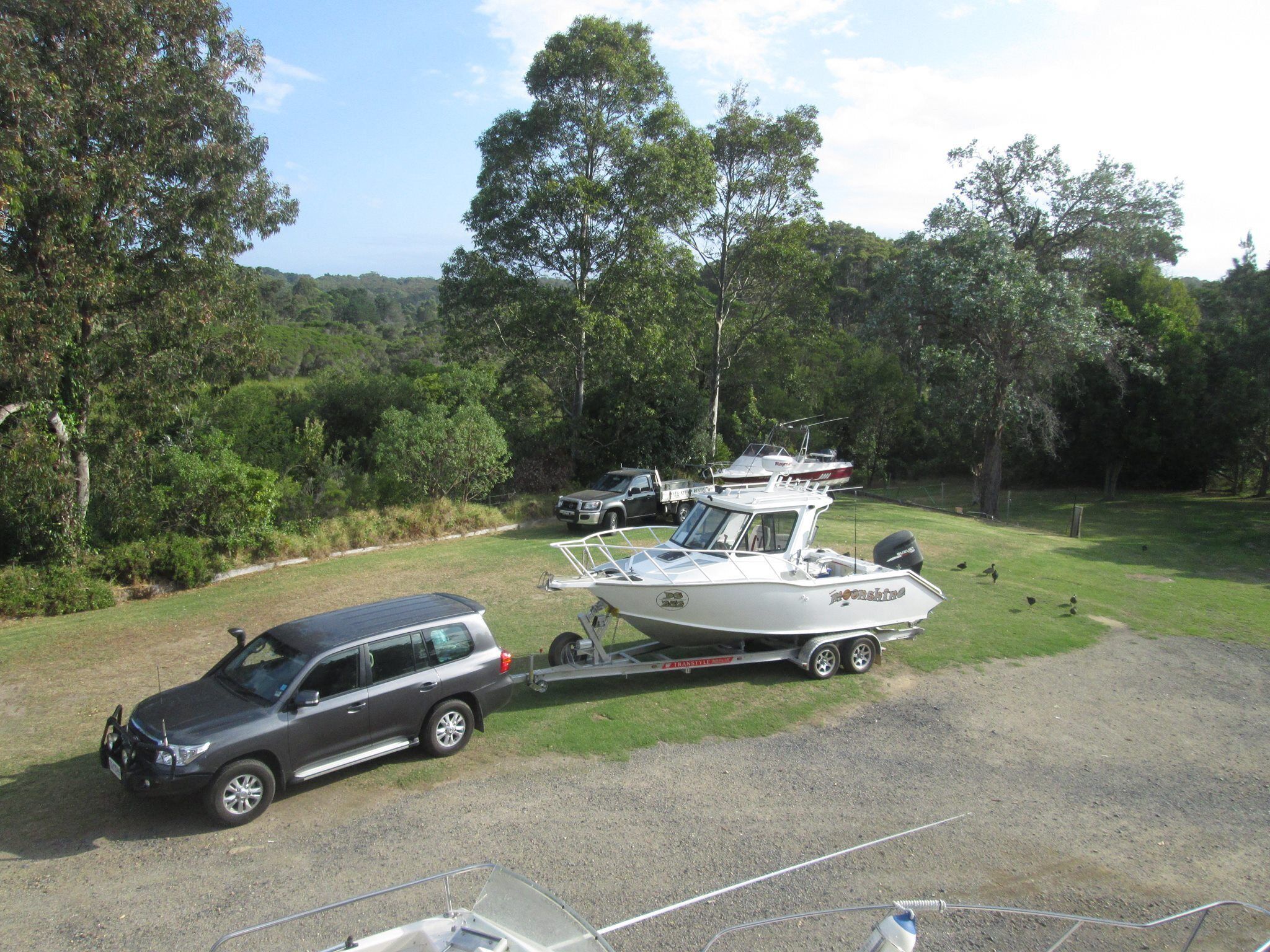 A boat trailer with a boat and a vehicle parked in a grassy area with trees in the background.