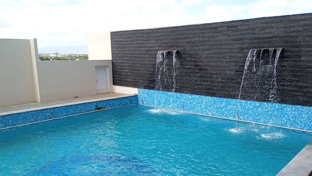 A swimming pool with clear blue water, a tiled edge, and a black textured wall with two waterfalls. - KRV Meridian (Modakurichi)