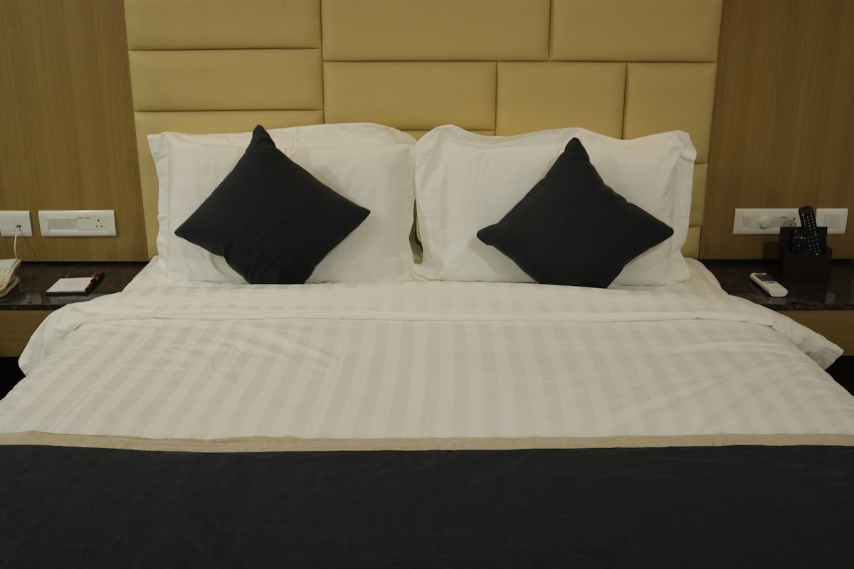 A hotel room with a bed, two black pillows, a bedside table with a phone and remote controls, and a wall socket.