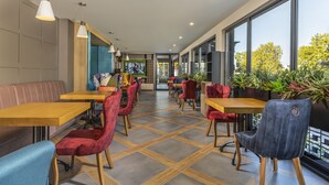 A modern restaurant interior with wooden tables and chairs, large windows, and indoor plants. - Urban Hotel (Plovdiv)