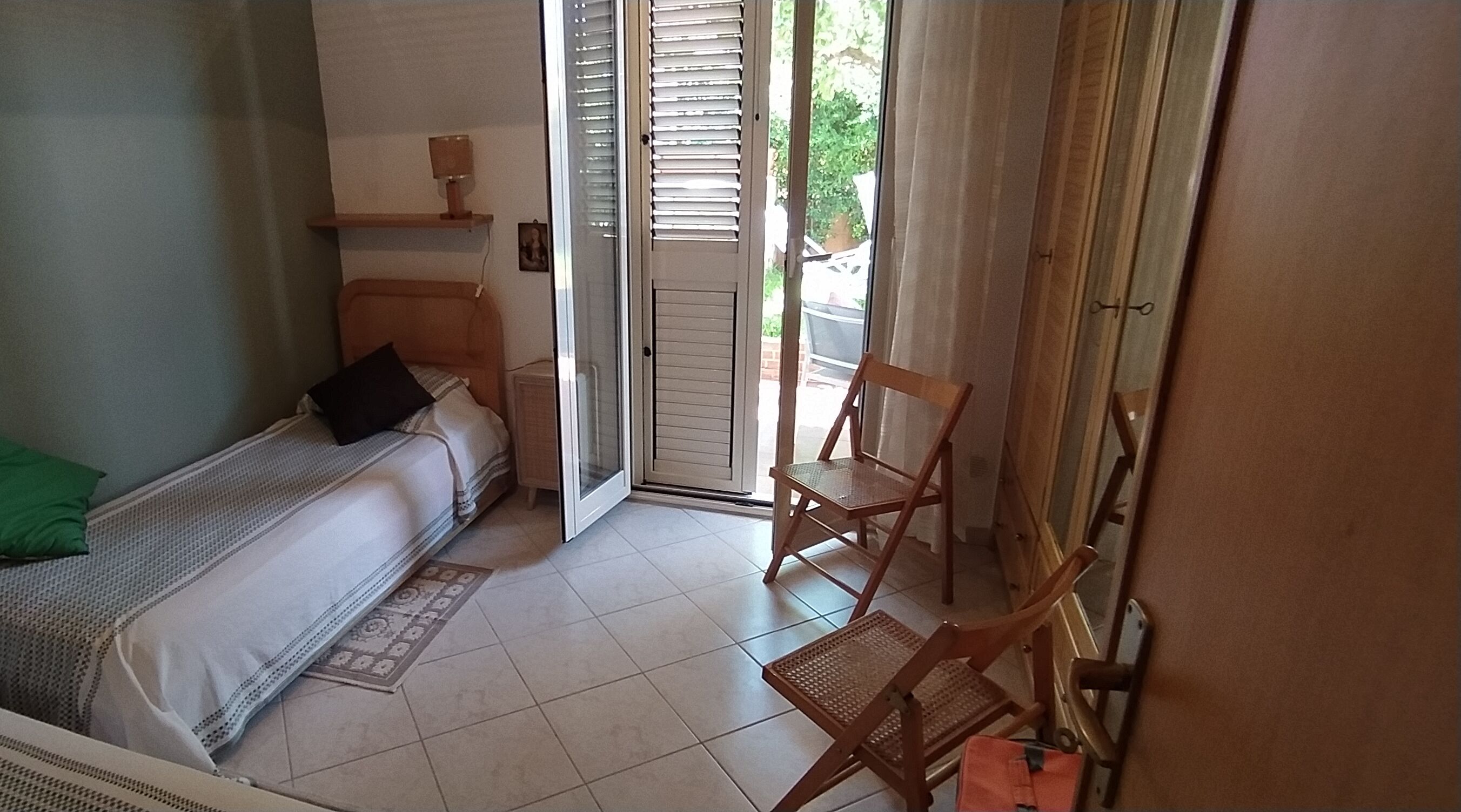 Ein Schlafzimmer mit Bett, Stuhl, kleinem Tisch und einer Tür zu einem Außenbereich.