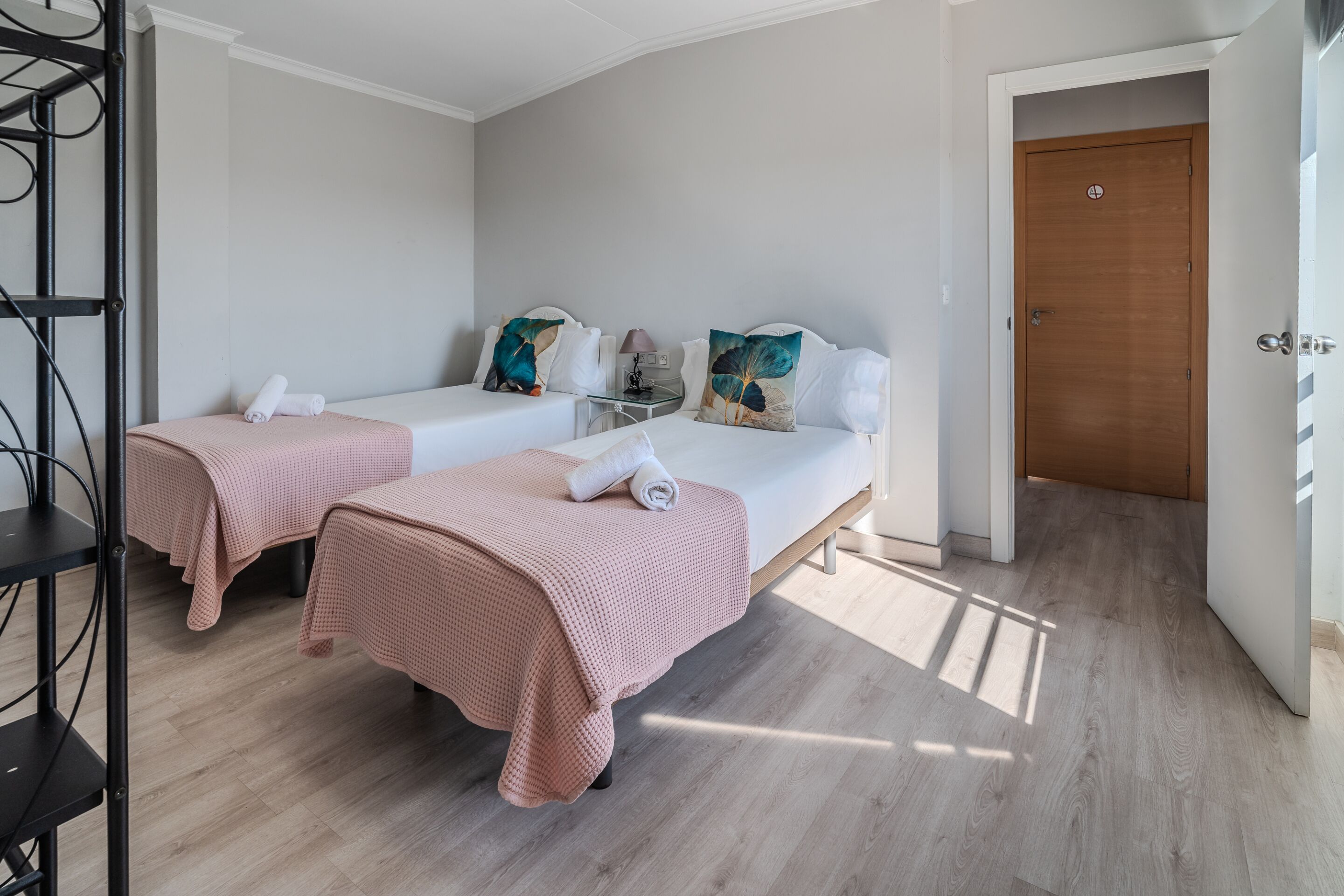 Una habitación con dos camas, cada una con una manta y almohadas rosas, una mesita con una lámpara y una puerta de madera.