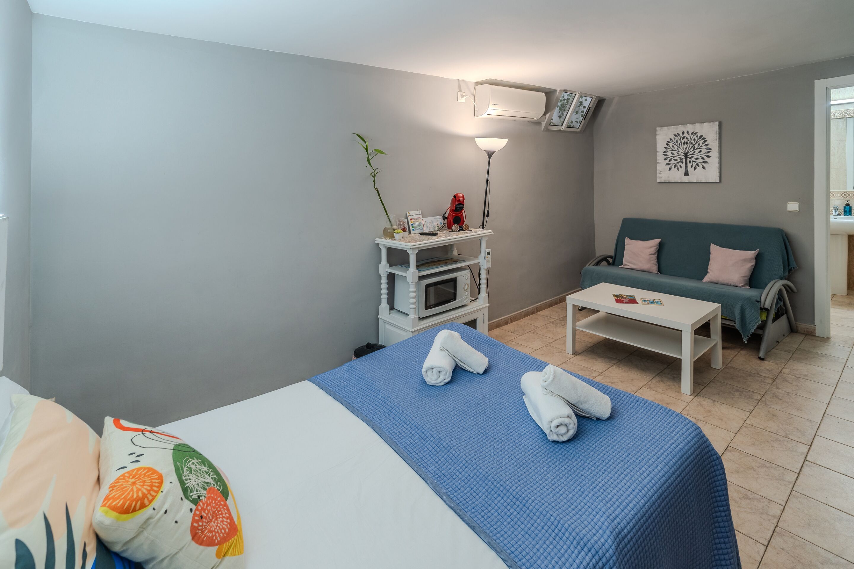 Une chambre d’hôtel avec un lit, une petite table blanche, un canapé, un four à micro-ondes et un climatiseur fixé au mur.