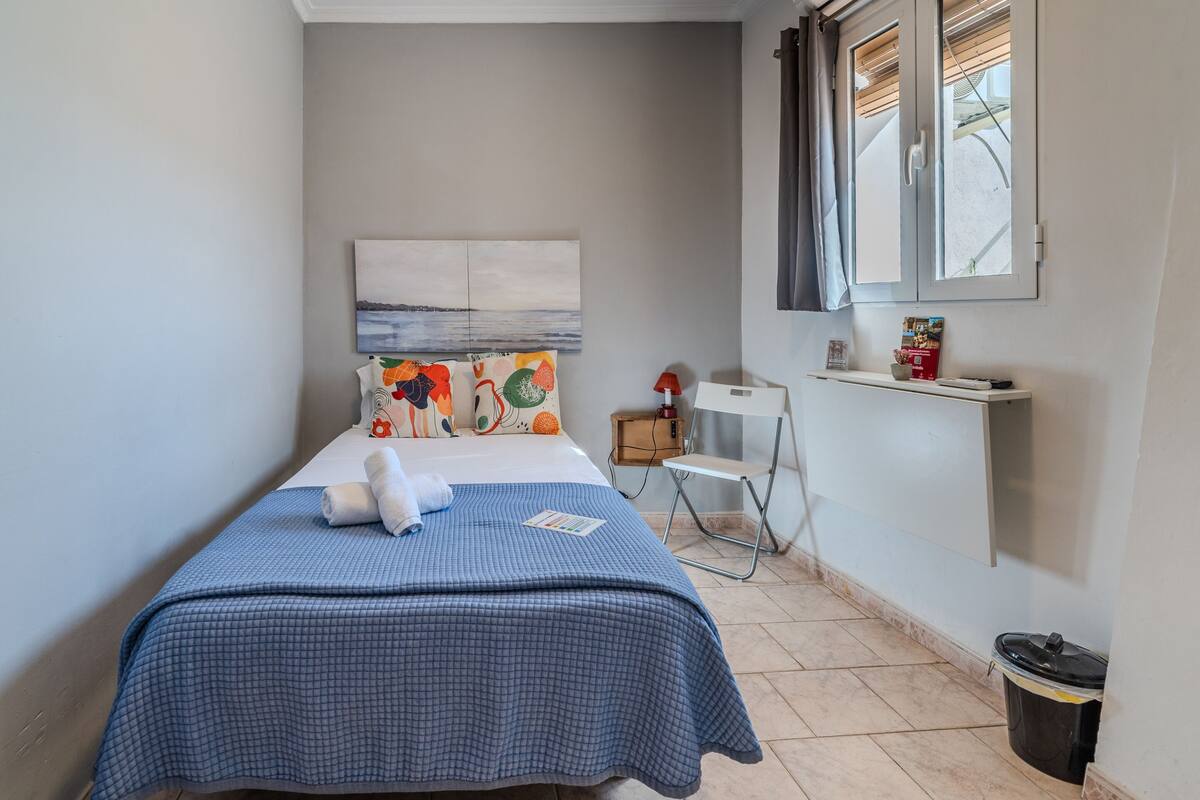 Ein ordentlich bezogenes Bett mit einer blauen karierten Tagesdecke, einem weißen Kissen und einem farbenfrohen Kissen mit Fruchtmotiven. Ein kleiner weißer Stuhl und ein Regal mit einem Buch und einer Schachtel stehen daneben.