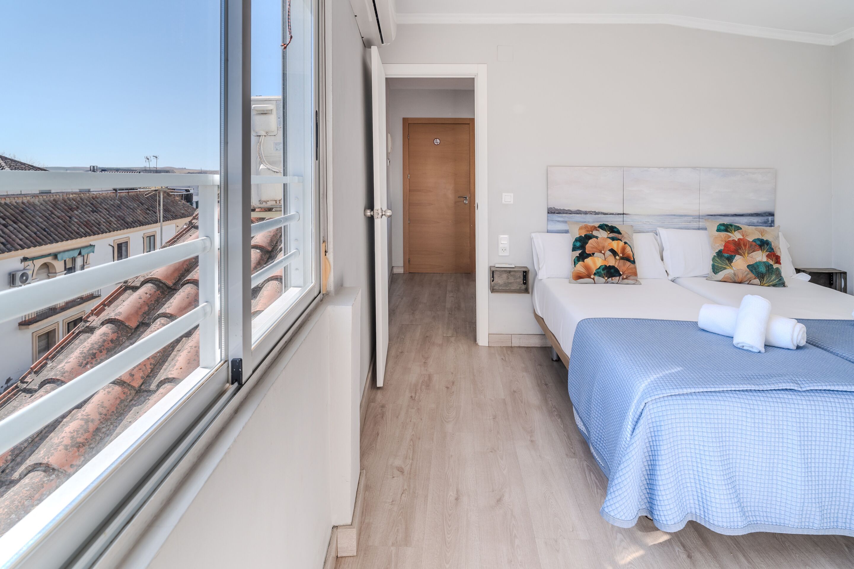Un dormitorio con una ventana grande, una cama con colcha azul a cuadros y vistas a edificios a través de la ventana.