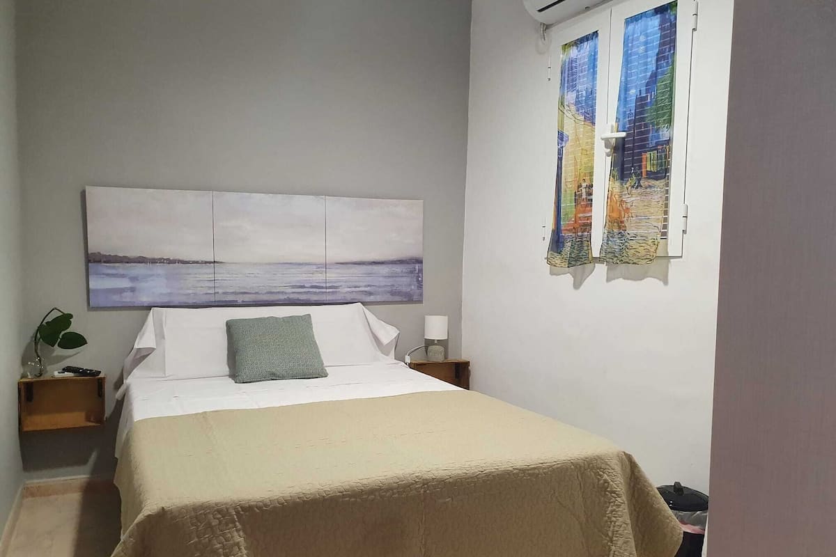 Ein Schlafzimmer mit Bett, Nachttisch, Wandbild, Fenster mit Blick und einer Pflanze.