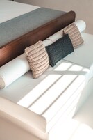 Una cama con cabecera de madera, dos mantas enrolladas y dos almohadas tejidas.