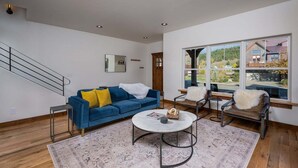A modern living room with a blue sofa, a round coffee table, and a large window offering a view of a house and greenery. - Downtown Location, Walk to Town, Close to River, AC (Pagosa Springs)