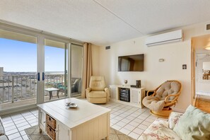 Un balcón con vistas a un paisaje urbano, un mueble blanco para televisor con una pantalla plana, un sillón beige y una silla de mimbre.