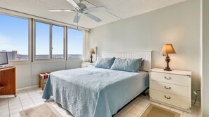 Een slaapkamer met een groot bed, een plafondventilator, een nachtkastje met een lamp, een bureau met een computer en uitzicht op de stad door grote ramen.
