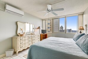 Un dormitorio con cama, cómoda, espejo, ventilador de techo, televisor y ventanas amplias con vista a la ciudad.