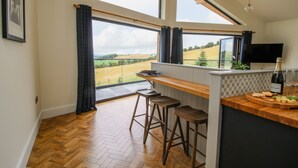 Eine Küche mit Holzboden im Fischgrätmuster, einer Bar mit Sitzgelegenheiten, einem großen Fenster mit Blick auf ein Feld und einer Champagnerflasche auf der Arbeitsfläche.