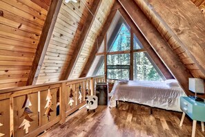 A bedroom with a sloped ceiling, a bed, a nightstand, a window, and a wooden railing with tree decorations. - Reindeer A Frame Cabin, hot tub, pet friendly (South Lake Tahoe)