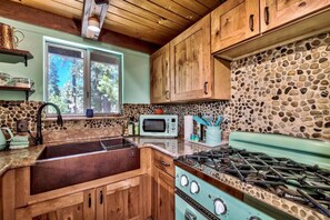 A rustic kitchen with wooden cabinets, a stone backsplash, a vintage stove, and a window with a view of trees. - Reindeer A Frame Cabin, hot tub, pet friendly (South Lake Tahoe)