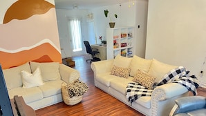 Una sala de estar con dos sofás, una mesa de centro y un mueble para televisión. Se observa una estantería con diversos objetos y una planta en maceta.
