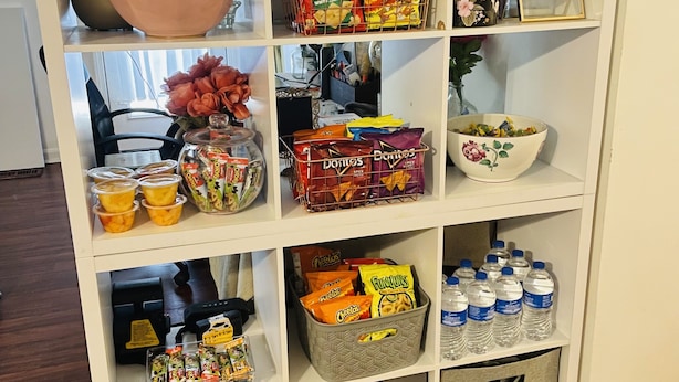 Un estante blanco con diversos productos de snack, como bolsas de papas y galletas, y un florero con flores.