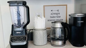 Una encimera de cocina con una licuadora Ninja, una cafetera Keurig y una jarra para café. Abajo, hay cajones con diferentes cápsulas de café.