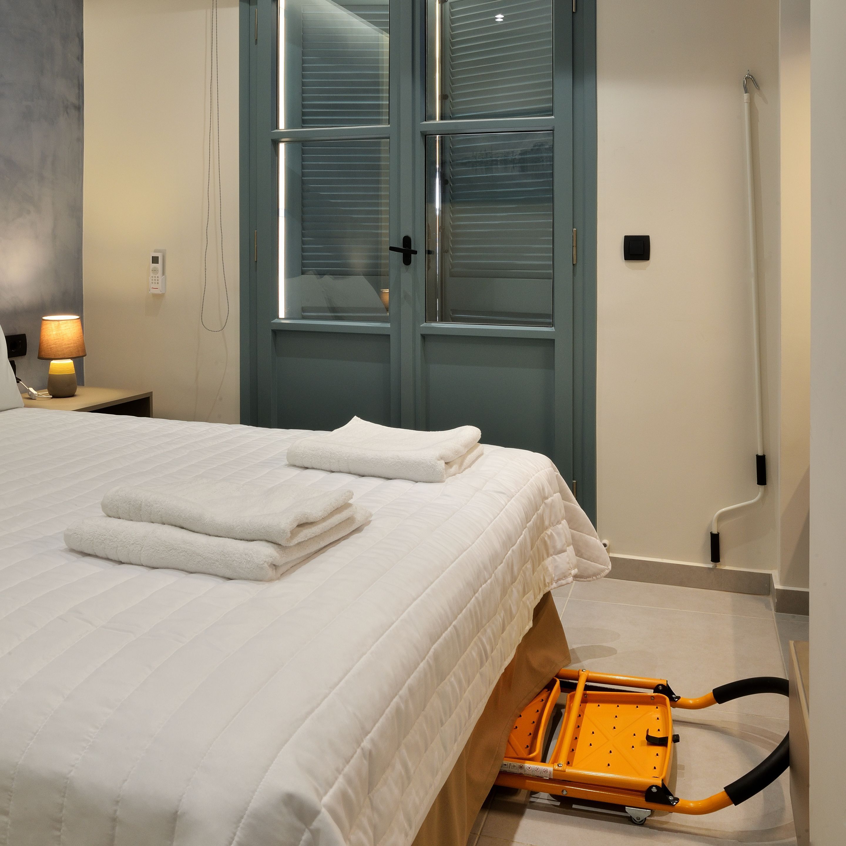 A neatly made bed with white linens and two folded towels, a bedside lamp, a phone, and a window with blinds.
