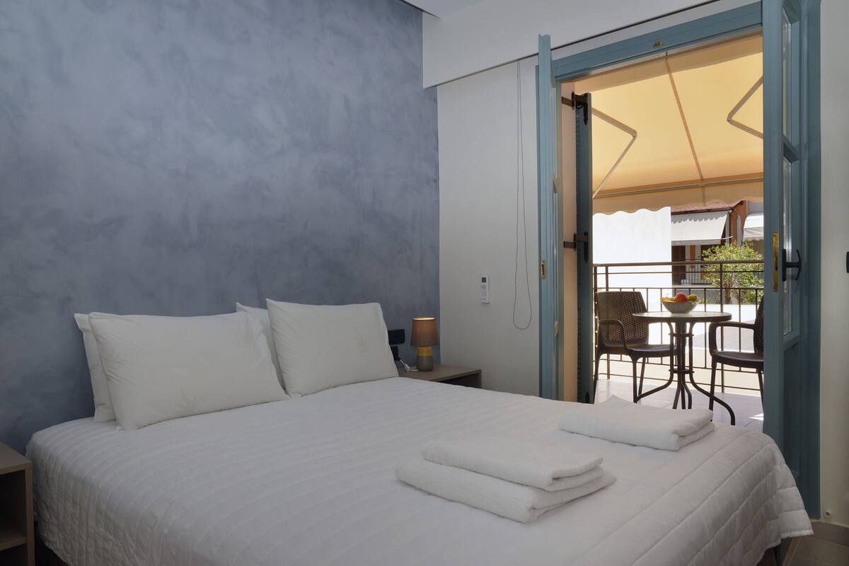 Un dormitorio con cama, almohadas, mesita de noche con lámpara, y puerta abierta que da a un área exterior con mesa y sillas.