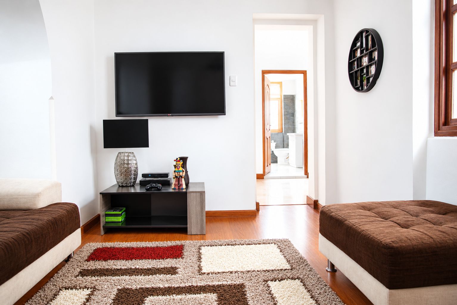 A modern living room with a flat-screen TV, a console table with a lamp and decorative items, two sofas, and a patterned rug.
