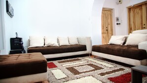 Quarto com sofá, um pufe, tapete estampado, portas de madeira e luminária fixada na parede.