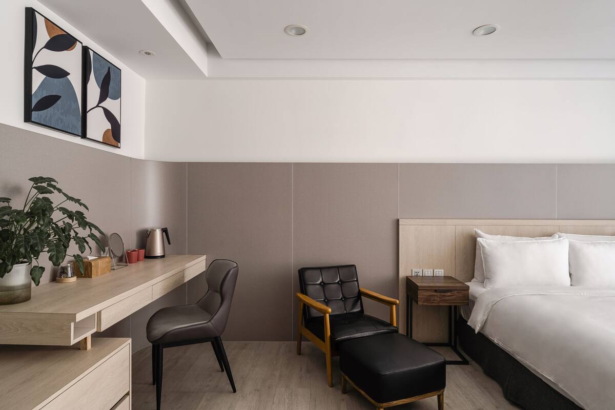 A modern hotel room with a wooden desk, a black leather chair, a bedside table with a lamp, and a large bed with white linens.