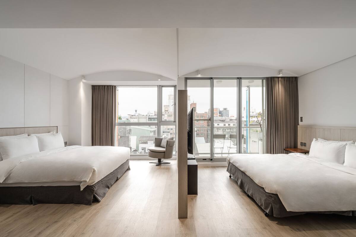 A modern hotel room with two beds, a sitting area, and large windows offering city views.