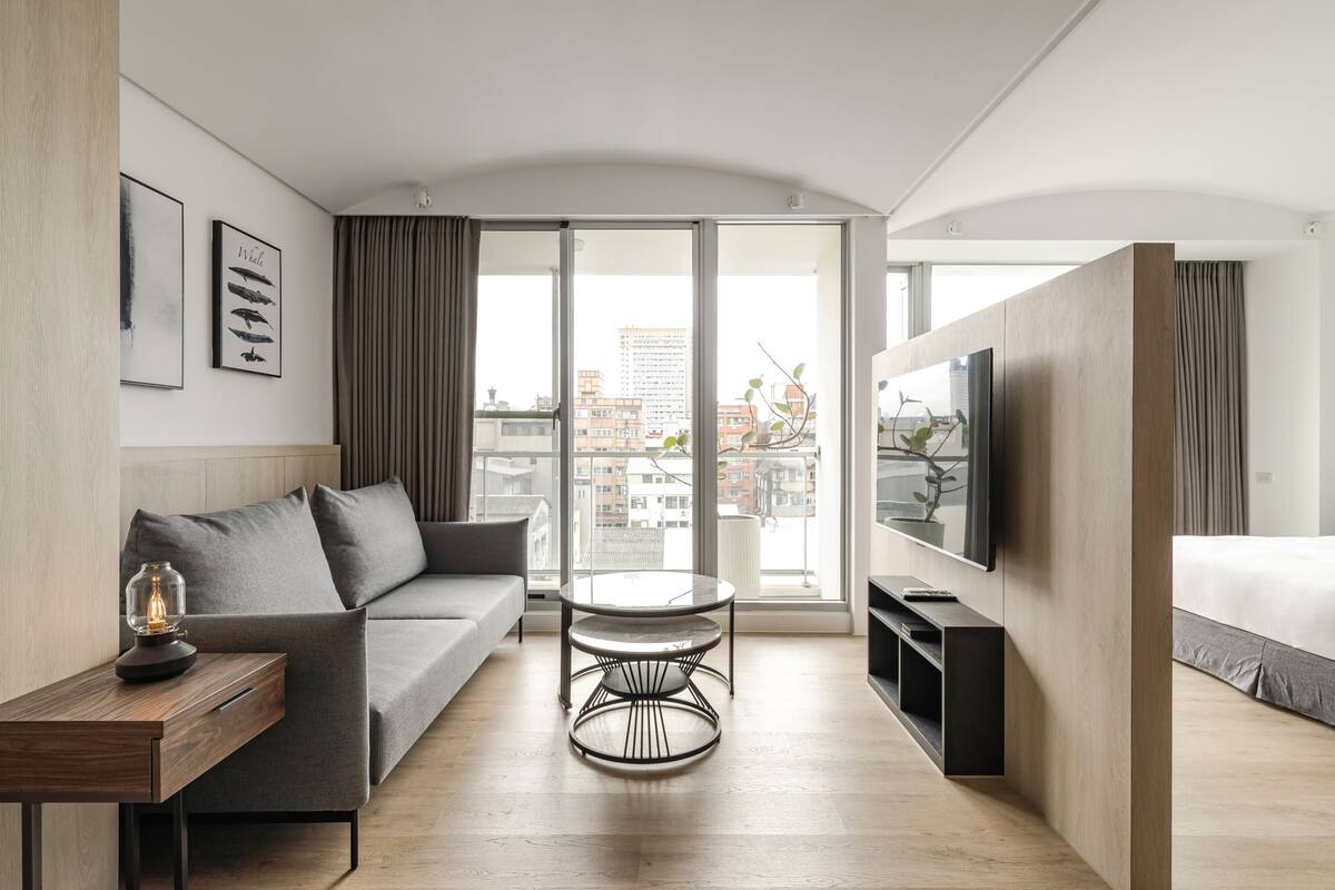 A modern hotel room with a sofa, a round table, a TV, and a balcony with a city view.