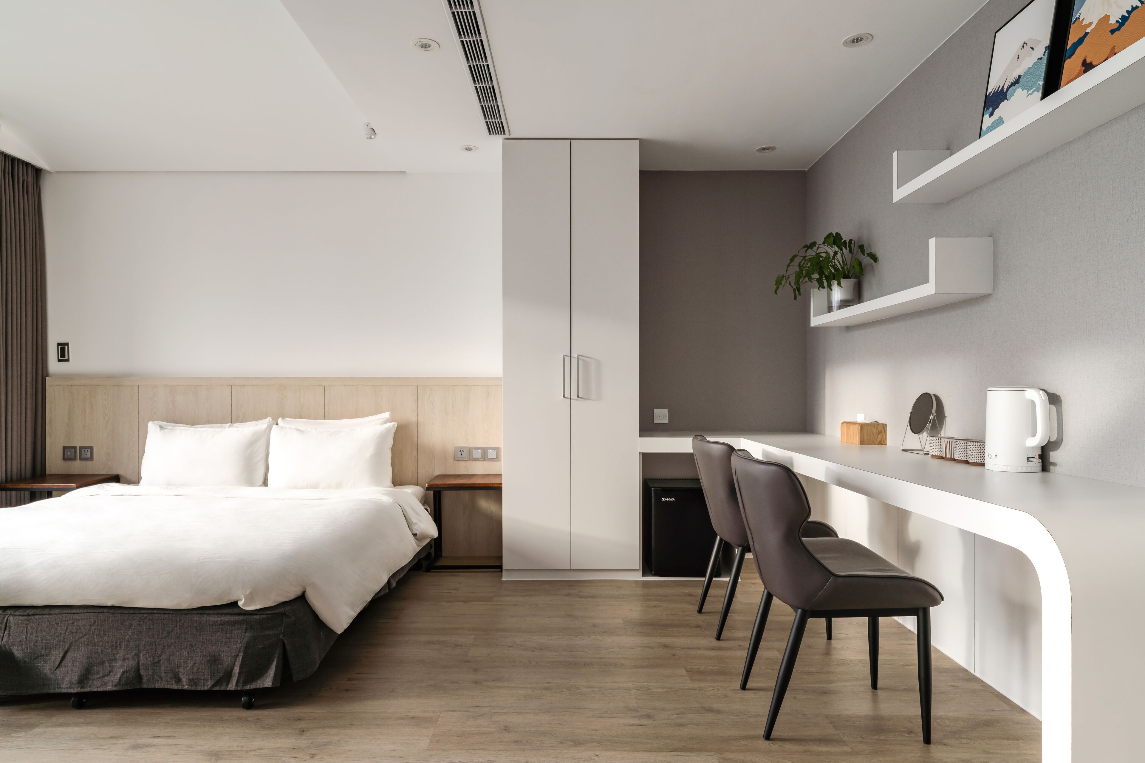 Una habitación de hotel moderna con cama, escritorio y sillas. Hay una pequeña kitchenette con cafetera y una planta en la estantería.