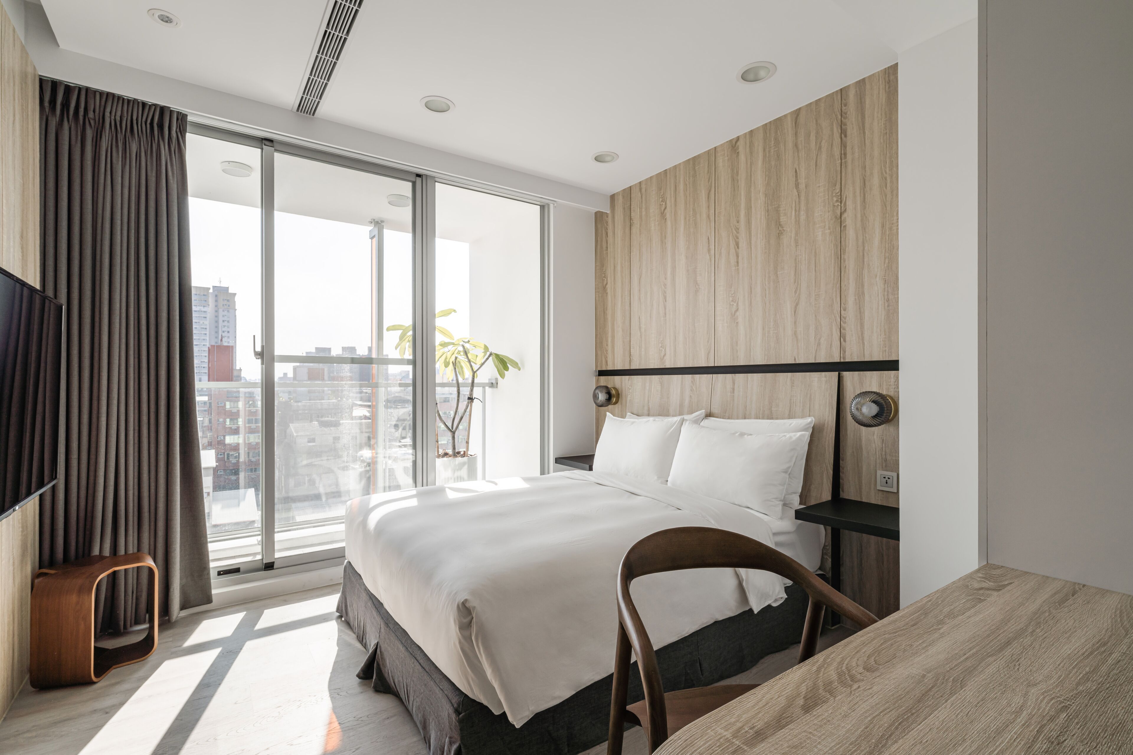 A modern hotel room with a large bed, a wooden headboard, a TV, a small wooden stool, and a view of the city through large windows.