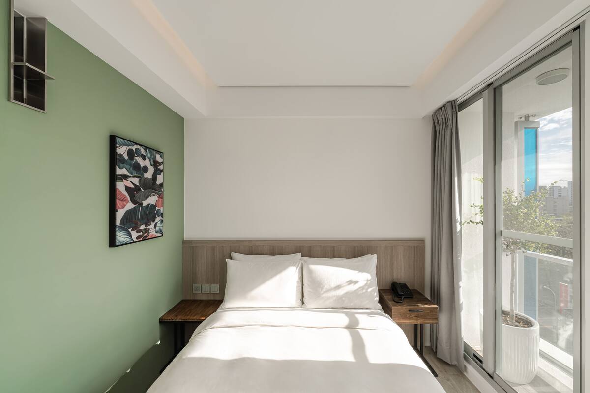 A modern hotel room with a large bed, a bedside table with a phone, a wall-mounted artwork, and a window with a view of the city.