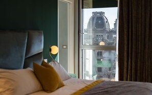 Un lit avec une tête de lit, des oreillers et une vue sur un immeuble par la fenêtre.