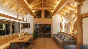 A spacious room with a high wooden ceiling, a dining area with a long table and chairs, a sofa, and a large wall-mounted artwork.