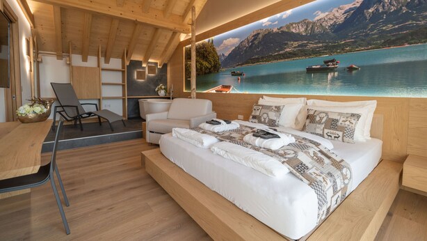 A modern bedroom with a large bed, a desk, and a view of a lake with boats.