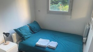 A bedroom with a bed, pillows, a nightstand, a lamp, and a window with a view of greenery. - Apartment near Paris (Noisy-le-Grand)